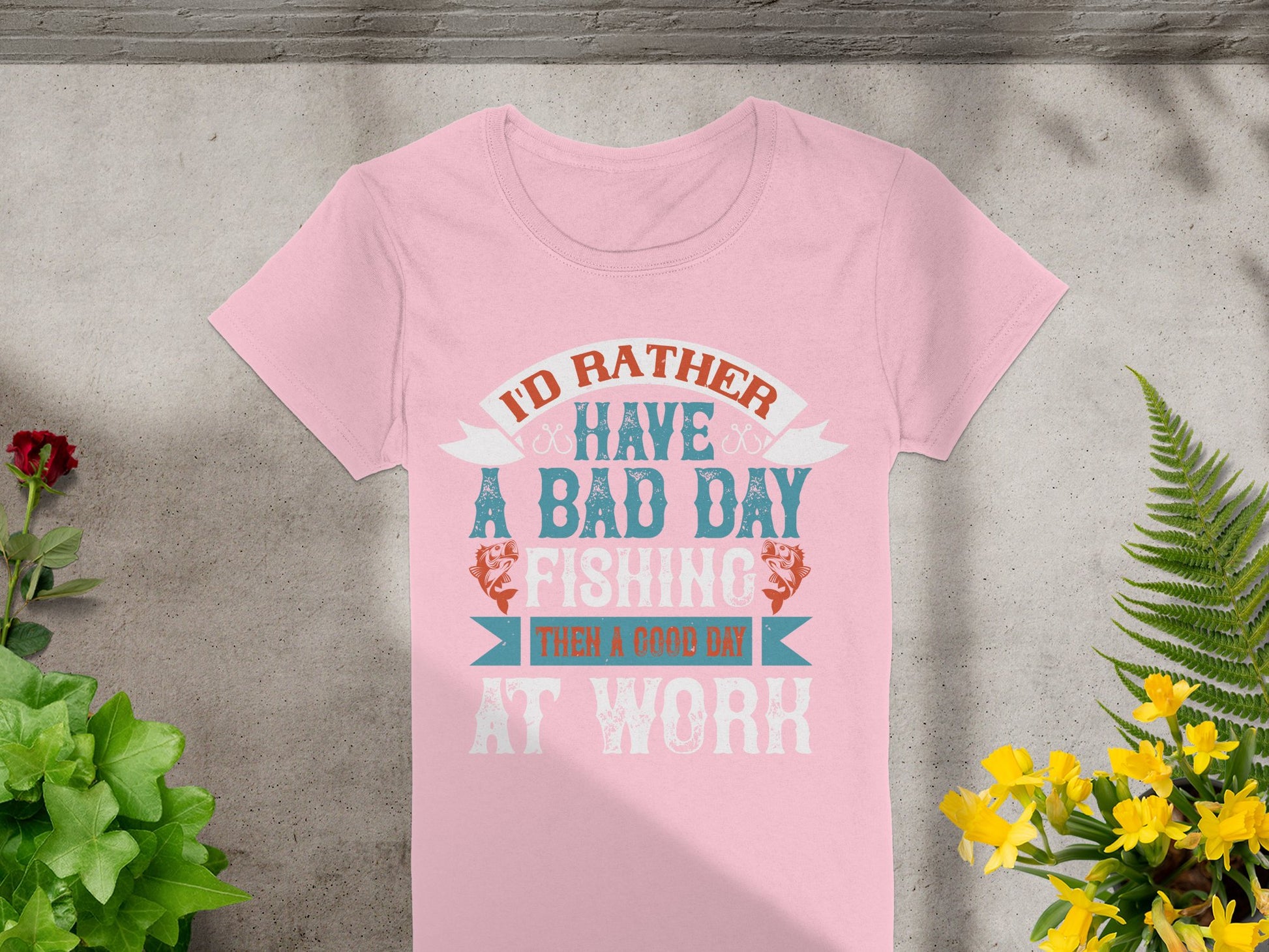 A pink Garment Graphics womens fishing T-shirt with the text Id rather have a bad day fishing than a good day at work is showcased on concrete, surrounded by vibrant green plants and yellow flowers, ideal for fishing enthusiasts.