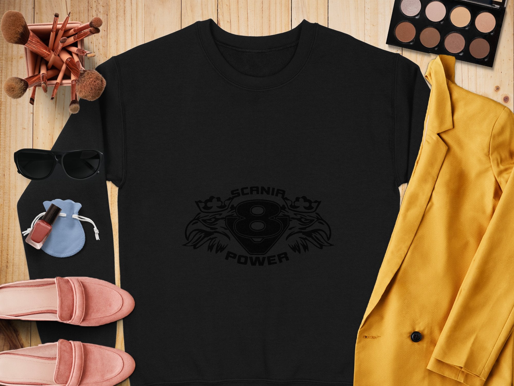 A black Garment Graphics Scania Power V8 sweatshirt with Scanir 8 Power design, paired with chic accessories: makeup brushes, sunglasses, a palette, lipstick, pink shoes, a blue pouch, and a yellow blazer—all on a wooden surface.