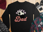 A black sweatshirt from Garment Graphics features a retro Soccer Dad design with Soccer above a ball and Dad in pink with stars. Its styled with a beanie, gloves, scarf, and plaid shirt on a wooden surface.