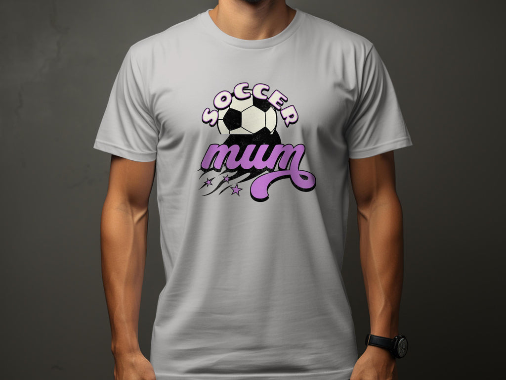 A person is wearing Garment Graphics classic fit light gray T-shirt with Soccer Mum in pink cursive, featuring a soccer ball and small stars on a plain dark background.