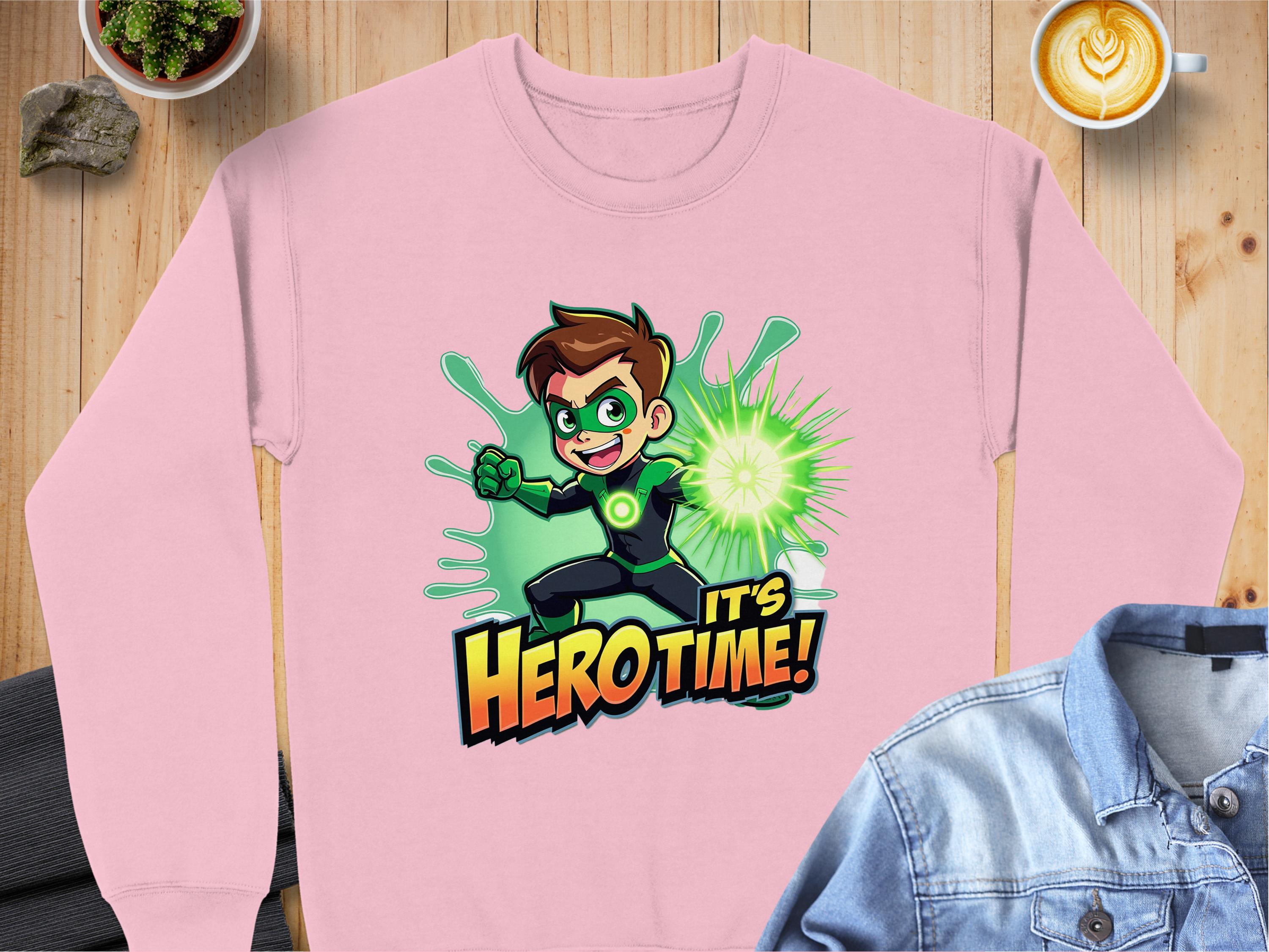 A pink Garment Graphics sweatshirt features a cartoon superhero boy in a green costume with a glowing orb. Ideal for adventurous youth, it’s styled on wood with rocks, a latte, and denim jacket to enhance its vibrant design.