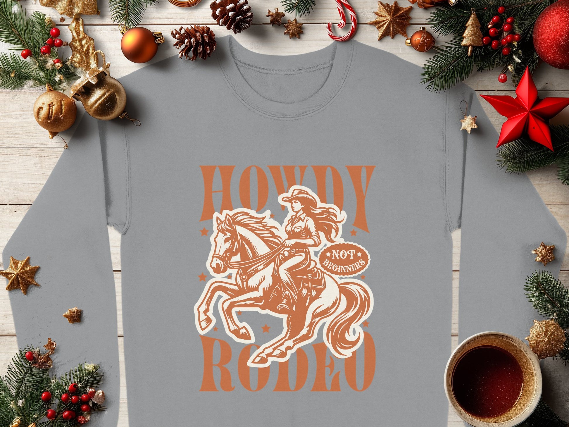 The Garment Graphics unisex gray sweatshirt features a rodeo cowgirl graphic with Howdy Rodeo above, adorned with pine branches, red stars, and candy canes—perfect for cozy coffee moments.