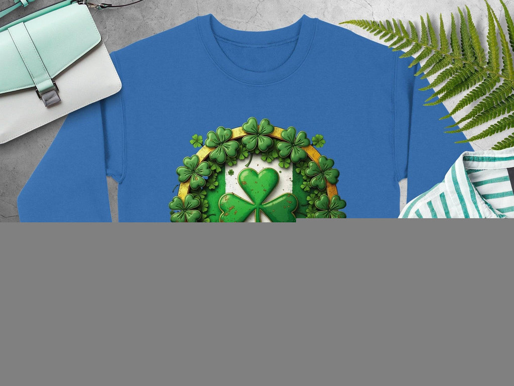 Blue Garment Graphics sweatshirt with a large shamrock, smaller shamrocks, and a central gold ribbon. Displayed flat on a gray surface with tan sandals, striped shirt, wristwatch, green bag, and ferns.