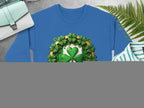 Blue Garment Graphics sweatshirt with a large shamrock, smaller shamrocks, and a central gold ribbon. Displayed flat on a gray surface with tan sandals, striped shirt, wristwatch, green bag, and ferns.
