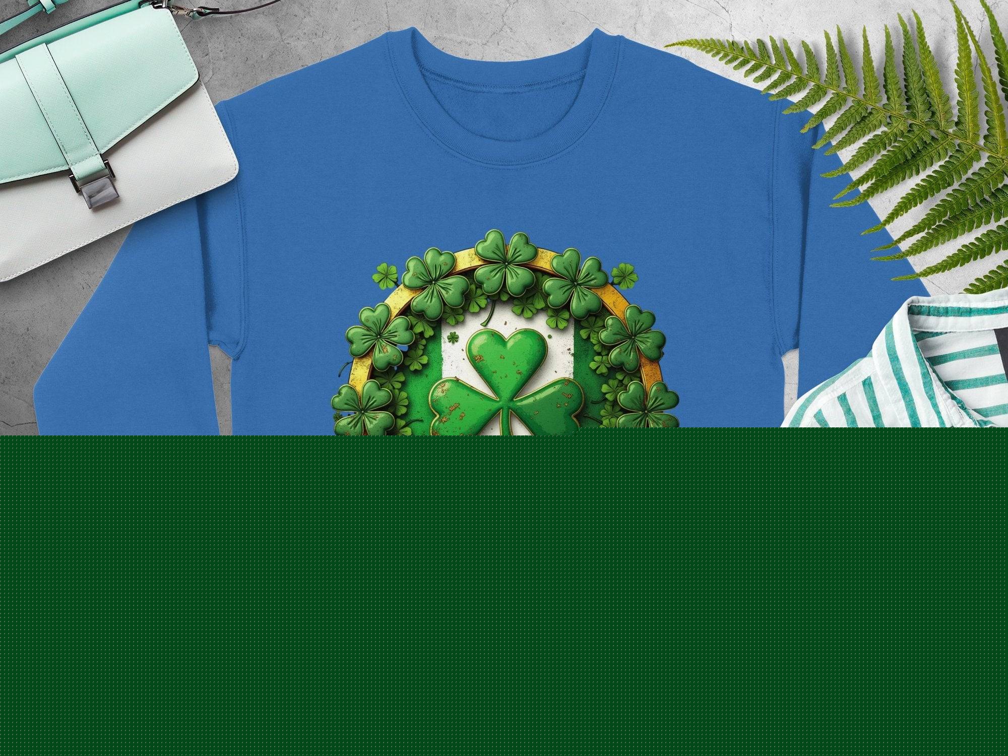 Blue Garment Graphics sweatshirt with a large shamrock, smaller shamrocks, and a central gold ribbon. Displayed flat on a gray surface with tan sandals, striped shirt, wristwatch, green bag, and ferns.