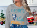 In a festive kitchen, someone holds a Santa-themed mug while wearing Garment Graphics gray DADDYS FISHING sweatshirt, featuring an illustration of a fisherman. Holiday decorations complete the cozy scene.