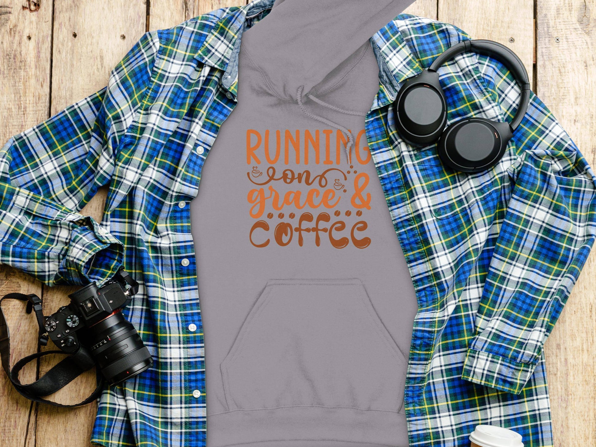 On a wooden surface, Garment Graphics presents a gray hoodie with Running on Grace & Coffee. Featuring a classic fit and crafted from medium-heavy fabric, its perfect for cozy days. Paired with a flannel shirt, headphones, camera, and coffee cup.