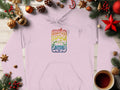 A light pink, retro graphic print hoodie by Garment Graphics featuring Woodstock 1969 and a car illustration is surrounded by holiday decorations, pine branches, ornaments, and a steaming cup of coffee on a wooden surface.