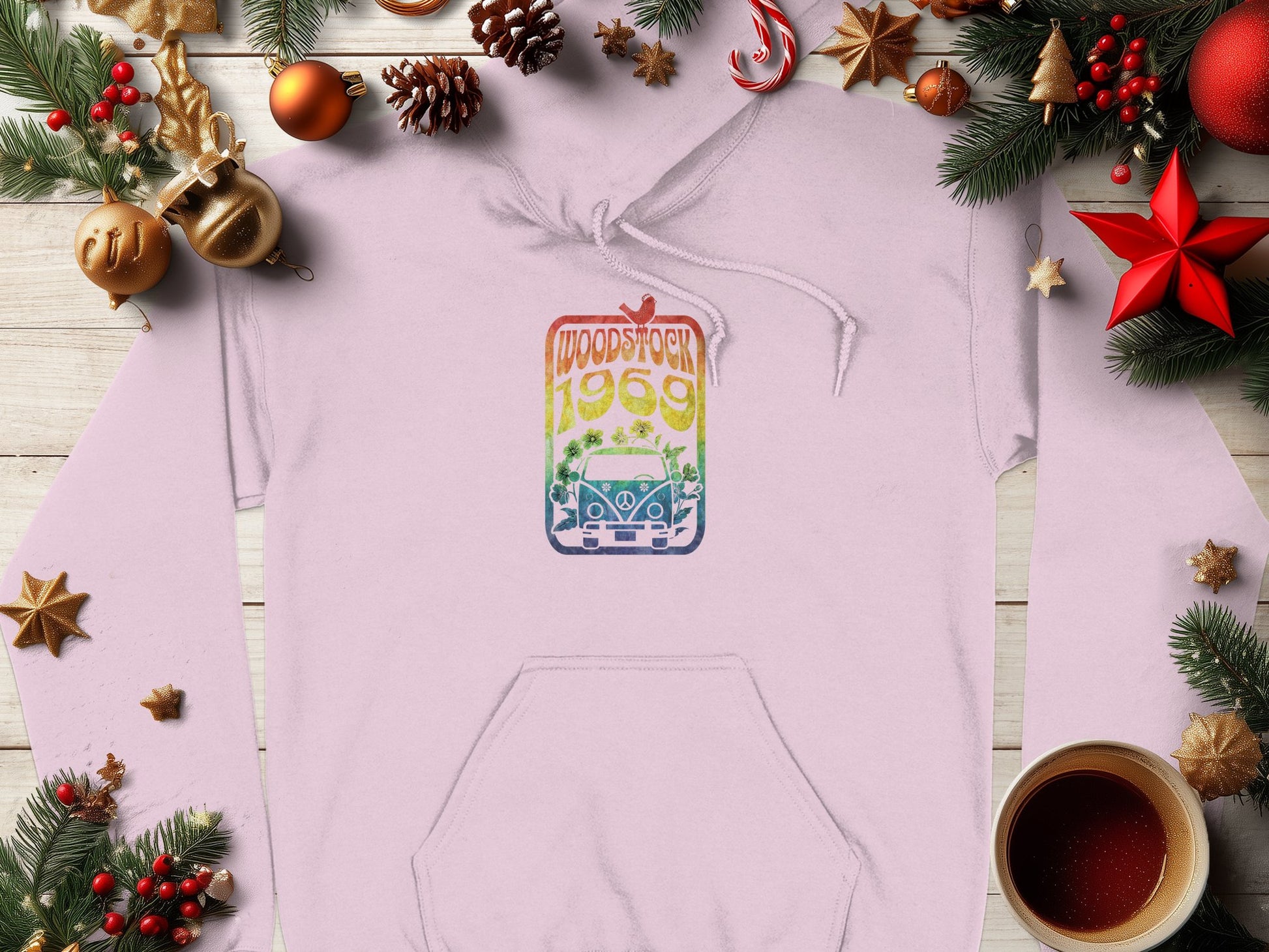 A light pink, retro graphic print hoodie by Garment Graphics featuring Woodstock 1969 and a car illustration is surrounded by holiday decorations, pine branches, ornaments, and a steaming cup of coffee on a wooden surface.