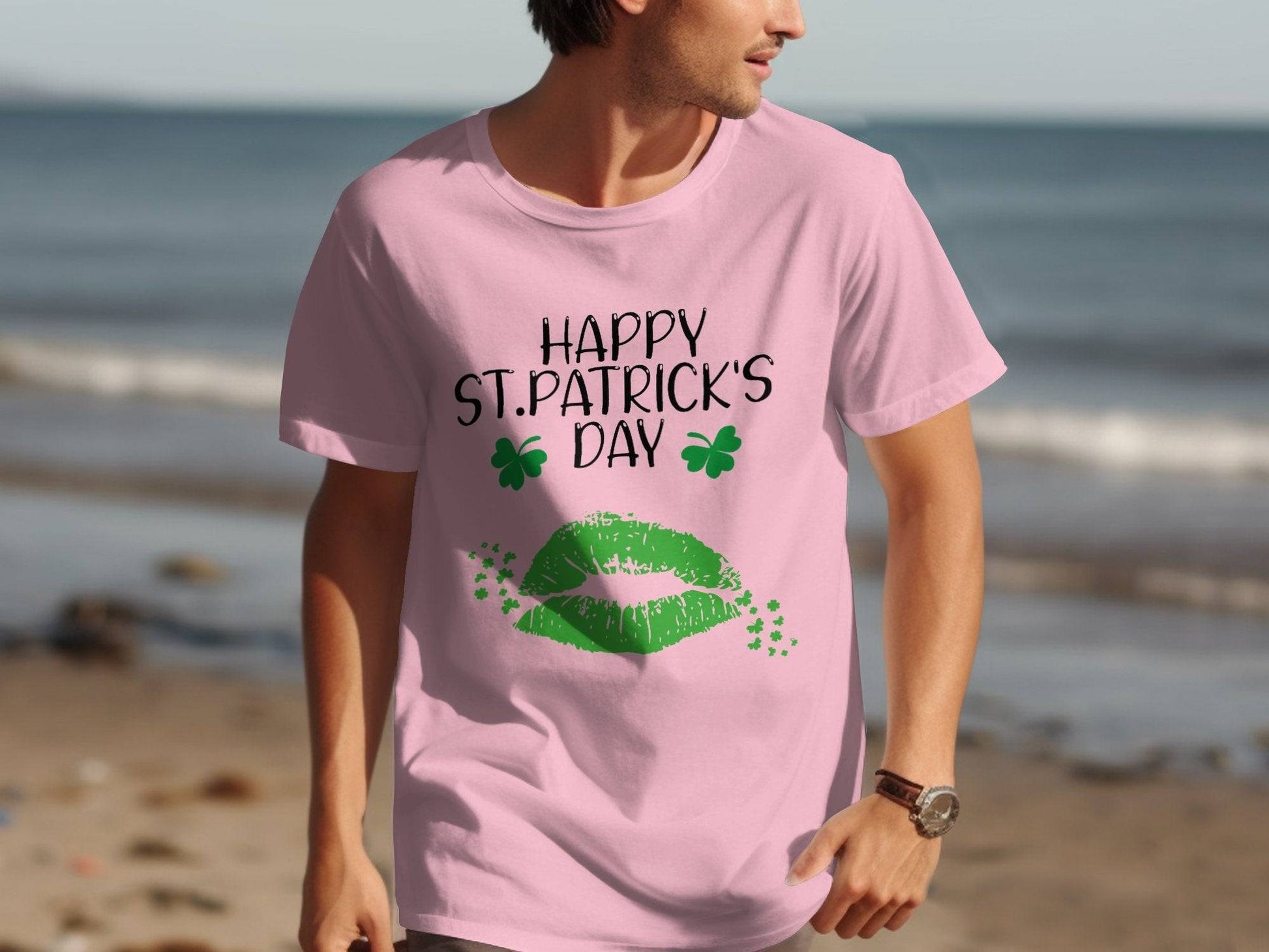 A man strolls by the beach in a vibrant pink Garment Graphics T-shirt with Happy St. Patricks Day, adorned with green lips and clovers, set against the serene sand and water backdrop.