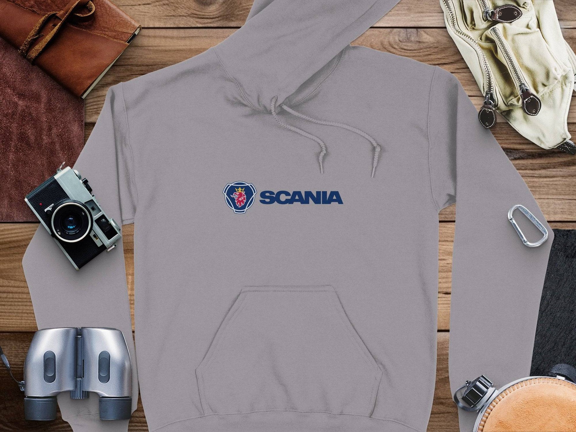 A gray Garment Graphics Scania hoodie, showcasing the iconic logo, lies on a wooden surface. Nearby are distinctive items: a vintage camera, binoculars, gloves, a carabiner, and a leather wallet.