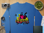 Garment Graphics blue sweatshirt features a vibrant graffiti design with a broken heart and I Love Graffiti in bold. Embrace urban style with nearby items like a camera, lens, kiwi bowl, map, notebook, and bag on a wooden surface.