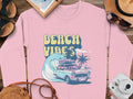 A pink Garment Graphics sweatshirt with a Beach Vibes design featuring a classic car, surfboards, waves, and palm trees lies on wood. A camera, notebook, hat, sunglasses, and sandals surround it to amplify the retro beach fun vibe.