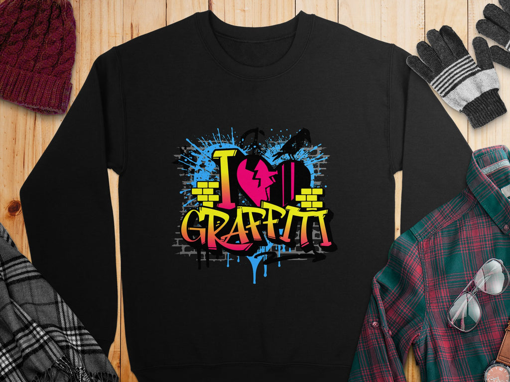 Garment Graphics presents a black sweatshirt with bold I Love Graffiti text in vivid graffiti-style letters. Splashes of blue and red paint over yellow brick wall motifs create an urban vibe, ideal with a hat, gloves, and plaid clothing on a wooden backdrop.