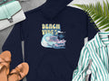 A navy hoodie from Garment Graphics features a surfboard, vintage car, palm tree, and ocean wave graphic. Beach Vibes is printed above. Embrace retro beach style: the hoodie is paired with accessories, sandals, and a striped shirt on a gray surface.