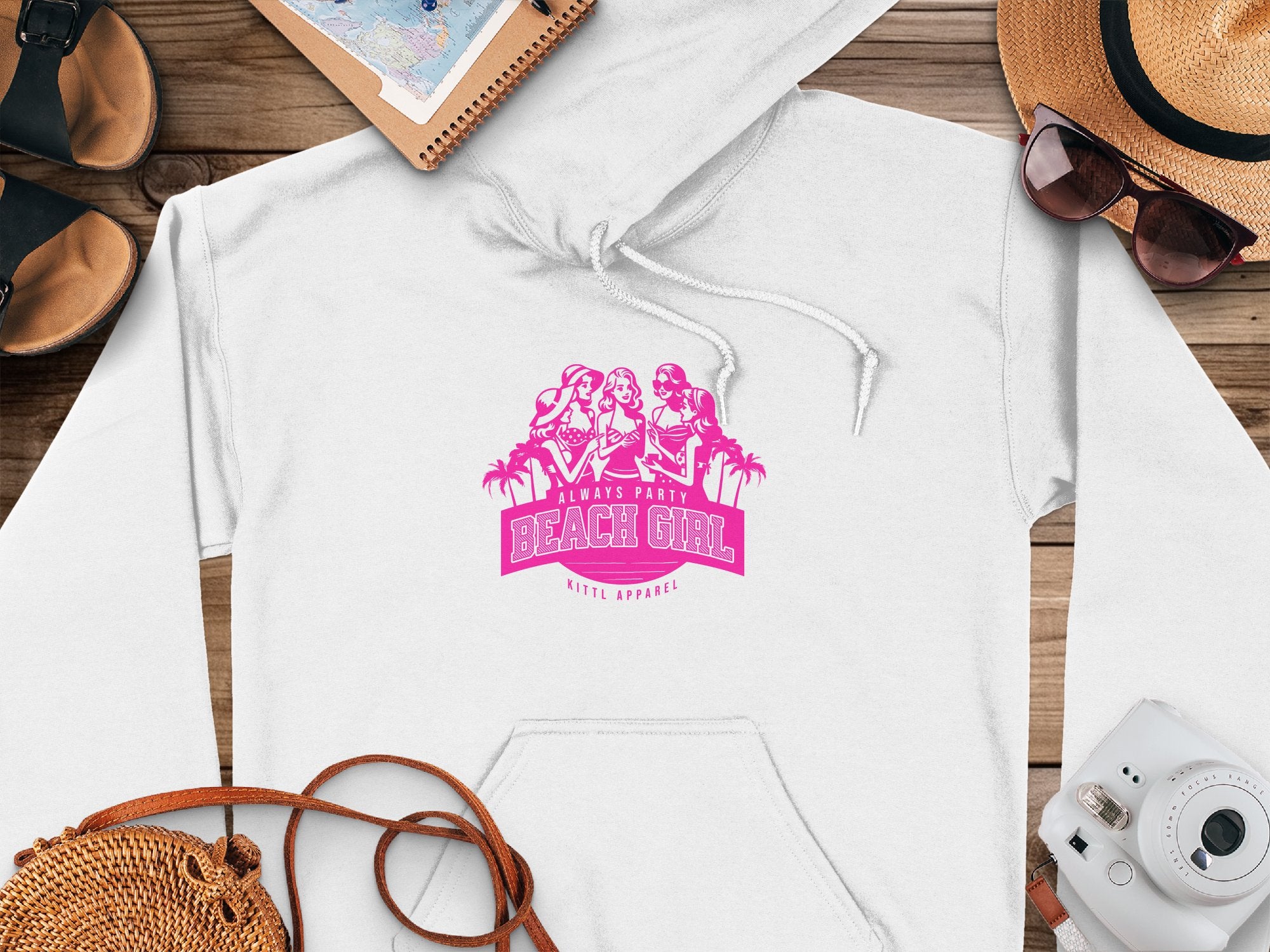 White hoodie from Garment Graphics features pink graphics of three cartoon women in beach attire with Always Party Beach Girl. Styled with a straw hat, sunglasses, sandals, and a camera on wood to complete the perfect casual look.