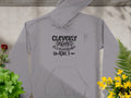 A gray hoodie from Garment Graphics, with text and decorative arrows, rests flat among lush green plants and vibrant yellow and red flowers on a concrete background—perfect for your casual wear collection.