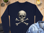 A navy Garment Graphics sweatshirt with an edgy style features a bold skull and crossbones on the front, lying on a wooden surface with a denim jacket, latte, and potted plant. Its unisex sizing for those who dare to stand out.