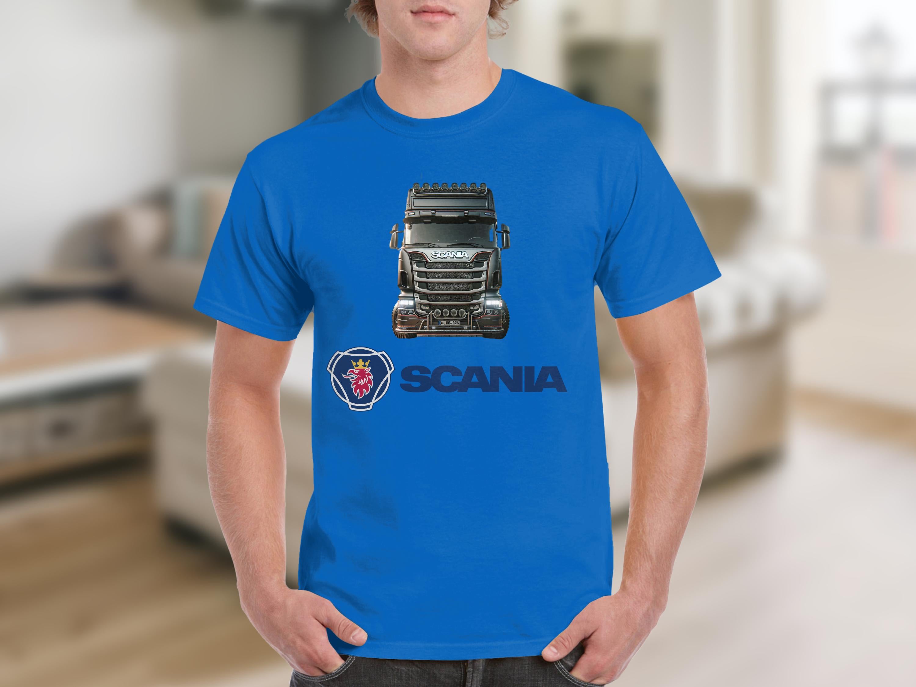 A person in a Garment Graphics blue T-shirt featuring a Scania truck graphic stands in a blurry room with a couch. Below the image, Scania and its logo are printed, ideal for truck enthusiasts.