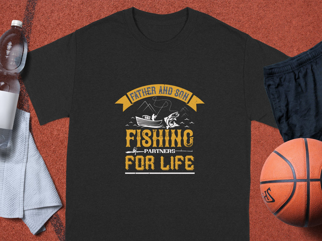 A black Garment Graphics Fishing T-Shirt displays Father and Son Fishing Partners for Life in yellow and white with a fish, rod, and boat design. Its paired with shorts, a basketball, towel, and water bottle on a brown surface—a perfect family bonding scene.