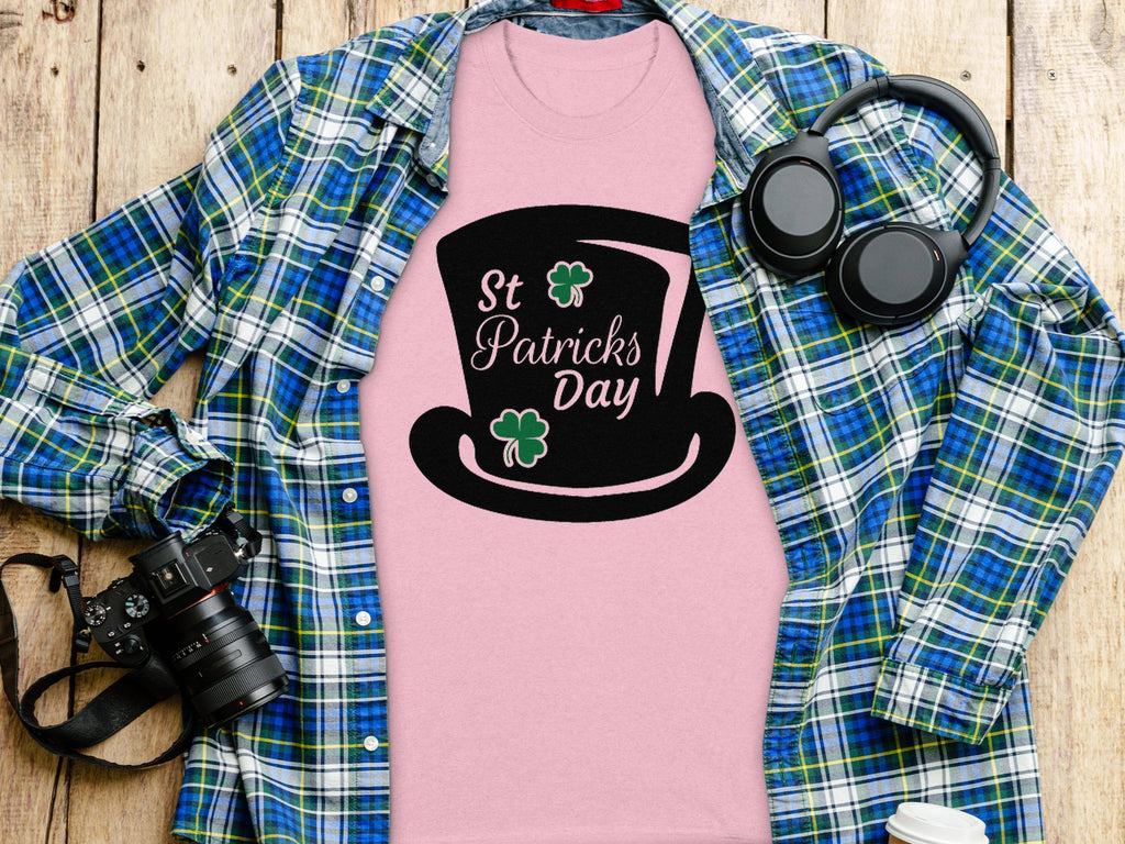 A pink Garment Graphics T-shirt with a black top hat and St Patricks Day text under a blue-green plaid shirt features whimsical four-leaf clover designs. Nearby, headphones, a camera, and a white paper cup adorn the wooden surface.