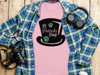 A pink Garment Graphics T-shirt with a black top hat and St Patricks Day text under a blue-green plaid shirt features whimsical four-leaf clover designs. Nearby, headphones, a camera, and a white paper cup adorn the wooden surface.