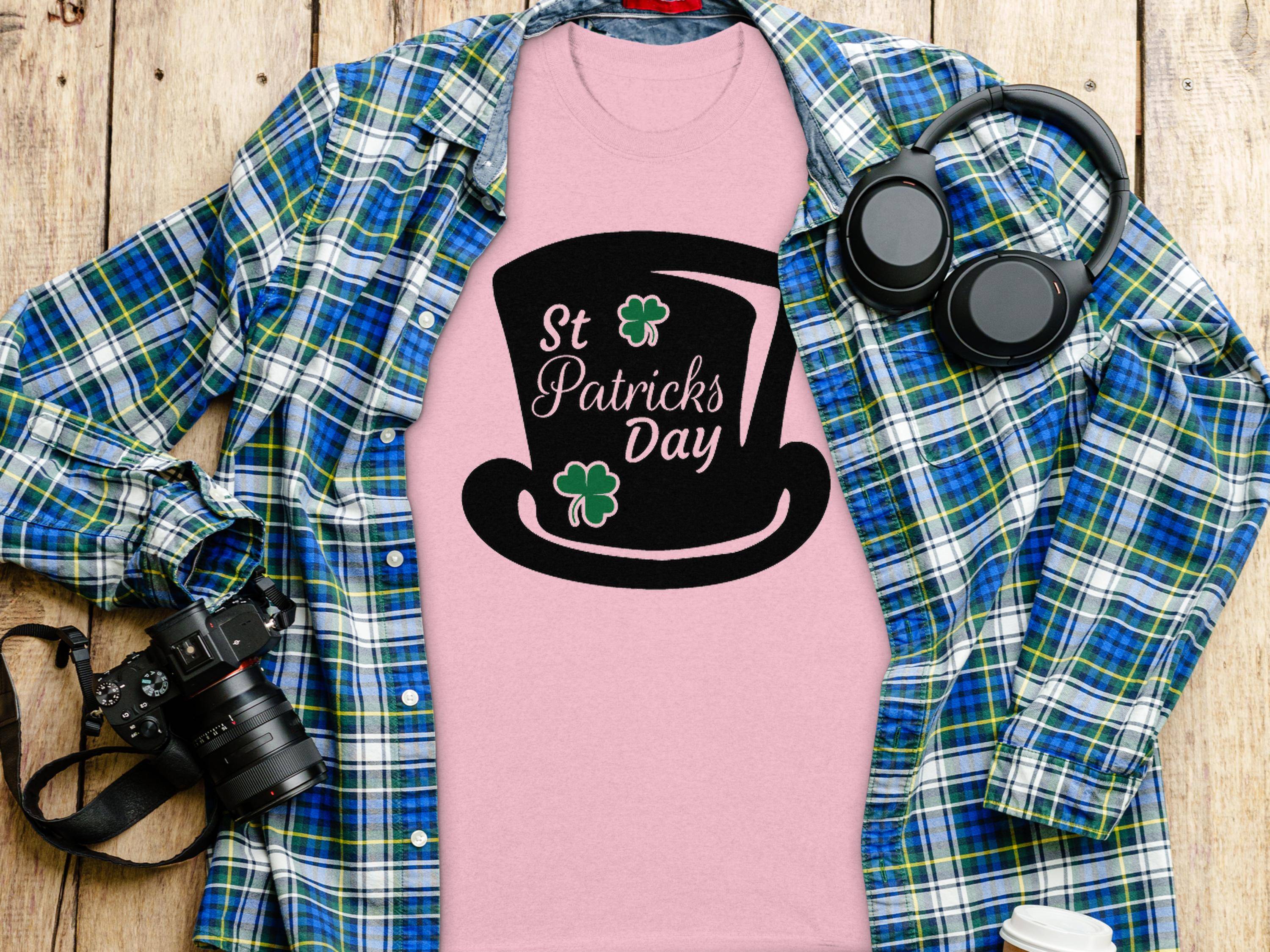 A pink Garment Graphics T-shirt with a black top hat and St Patricks Day text under a blue-green plaid shirt features whimsical four-leaf clover designs. Nearby, headphones, a camera, and a white paper cup adorn the wooden surface.