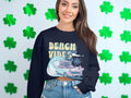 A woman with long brown hair sports a Garment Graphics navy sweatshirt that reads Beach Vibes with a retro design of a classic car, palm tree, and wave, standing against a backdrop adorned with green shamrocks.