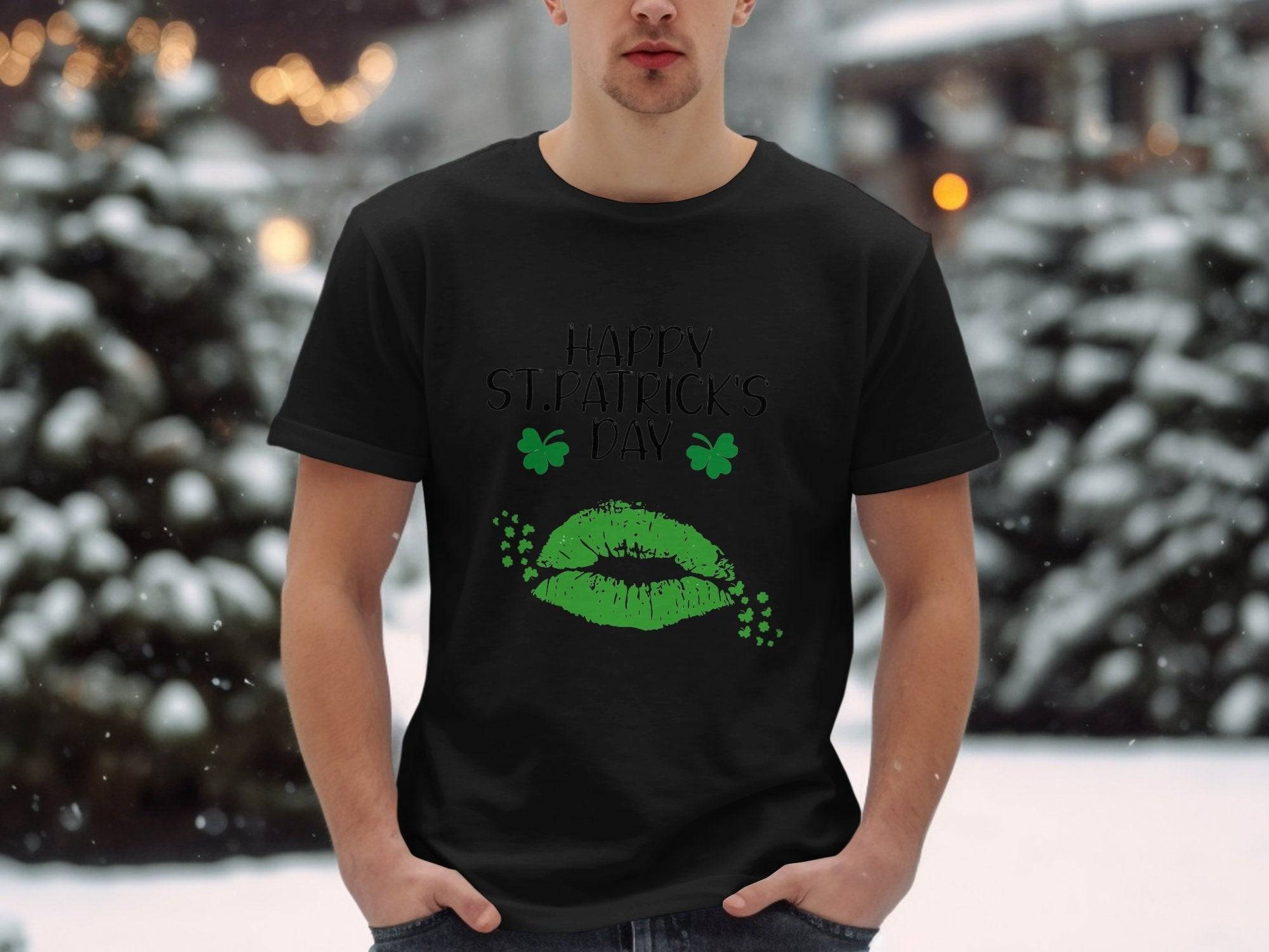 A person stands outside in the snowy landscape wearing a Garment Graphics T-shirt featuring festive green lips and clovers with the phrase Happy St. Patricks Day. Snow-laden trees and soft, blurred lights enhance the magical scene.