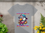 A Garment Graphics gray T-shirt with a unique design rests on concrete, featuring the Cool Brave Wild Swag Duck in glasses, a hat, and a jacket. The vibrant Ducks Dope text is framed by red and yellow flowers with green leaves.
