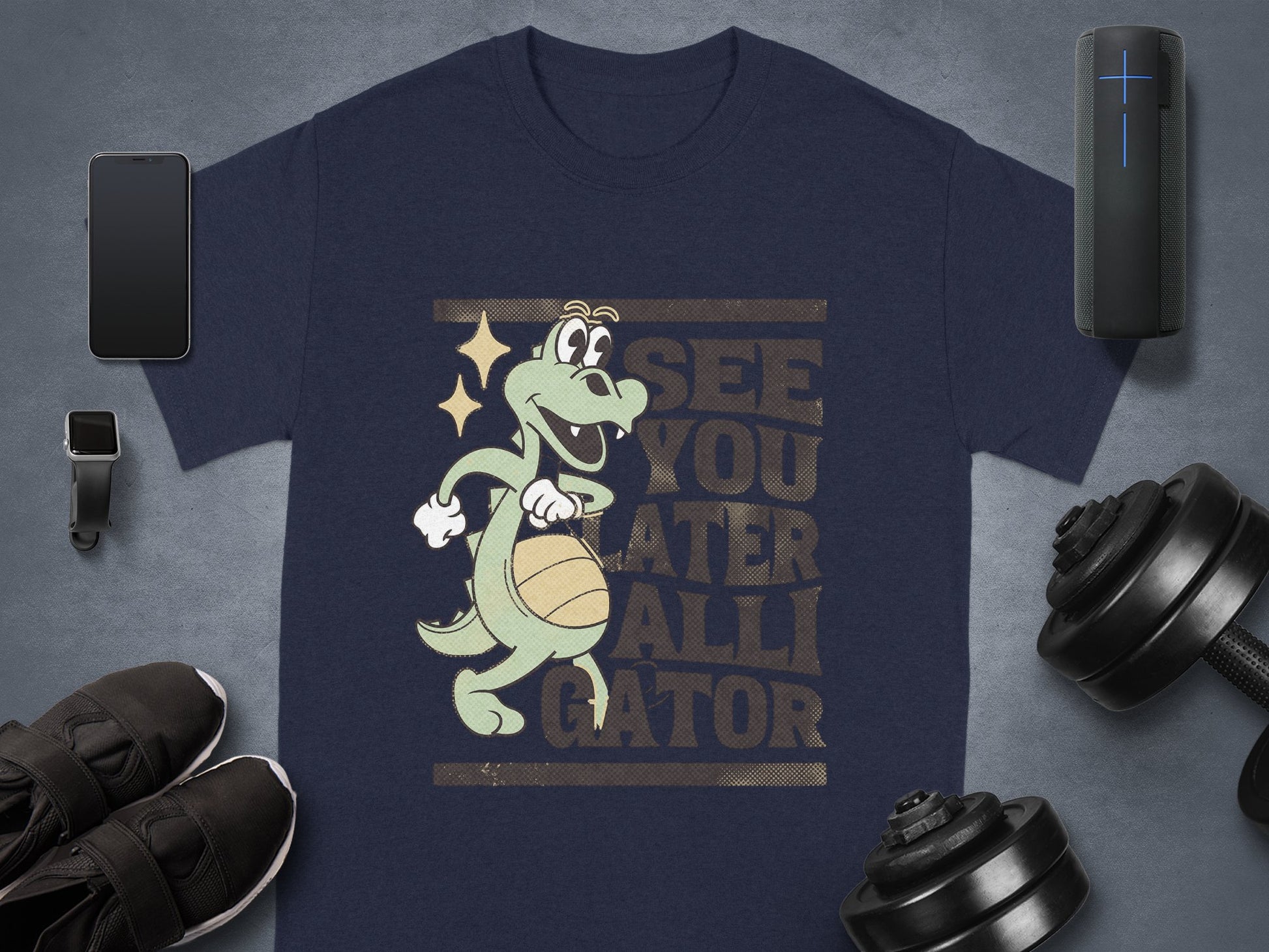Garment Graphics offers a navy blue t-shirt with a cartoon alligator spinning a basketball, displaying See You Later Alligator. Ideal for casual wear, its showcased on a gray background with a smartphone, smartwatch, sneakers, dumbbells, and a portable speaker.