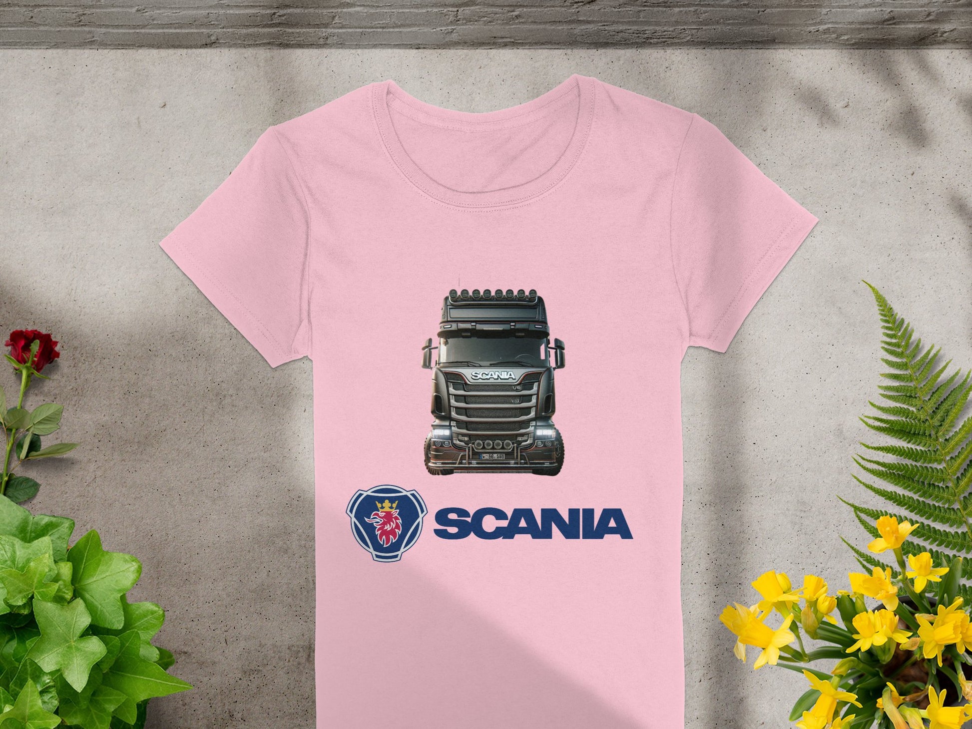 This pink Garment Graphics T-shirt is ideal for automotive enthusiasts, featuring a Scania truck graphic above the logo and name. The vibrant image is surrounded by green plants and yellow flowers on concrete, making it perfect for graphic T-shirt lovers.