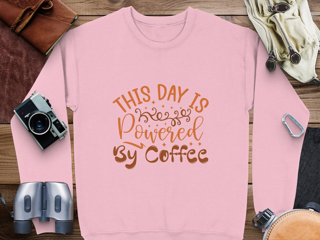Garment Graphics presents a pink graphic sweatshirt with This Day is Powered By Coffee in brown font. Surrounded by a camera, binoculars, leather bag, gloves, and climbing gear on wood, its ideal for adventurers fueled by caffeine.