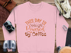 Garment Graphics presents a pink graphic sweatshirt with This Day is Powered By Coffee in brown font. Surrounded by a camera, binoculars, leather bag, gloves, and climbing gear on wood, its ideal for adventurers fueled by caffeine.