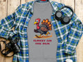 A flat lay displays a Garment Graphics gray T-shirt featuring a cartoon turkey with the caption Turkey on the Run, under a blue and green plaid shirt. It humorously sits next to headphones, a camera, and a coffee cup on wood.