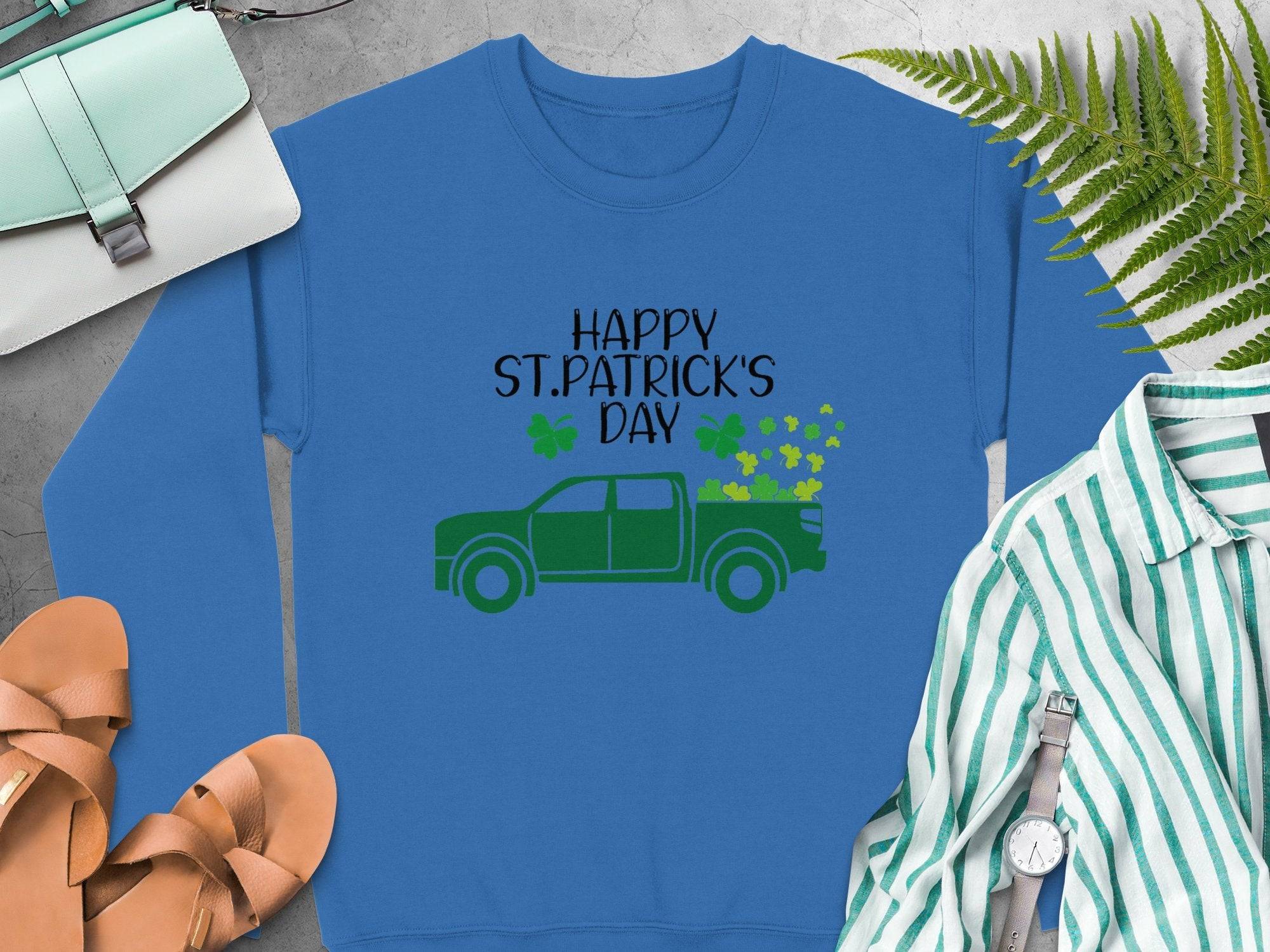 Garment Graphics festive sweatshirt displays a green truck with shamrocks and Happy St. Patricks Day text on blue. Styled flat, its paired with sandals, a striped shirt, watch, green purse, and ferns.