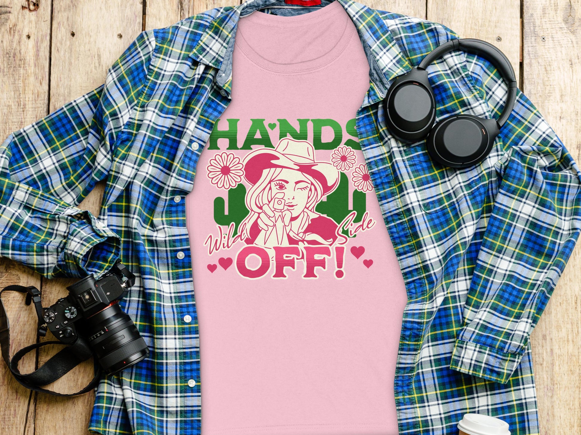 A Garment Graphics pink T-shirt with a bold cowboy-themed woman design and Hands Off! Wild Side text pairs stylishly with a blue-green plaid shirt, complemented by a camera and headphones on a wooden background.