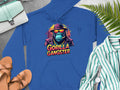 Streetwear style blue hoodie by Garment Graphics features a bold gorilla in a fedora and sunglasses with Gorilla Gangster text. Displayed on a gray surface with a purse, sandals, striped shirt, watch, and green fern leaves.