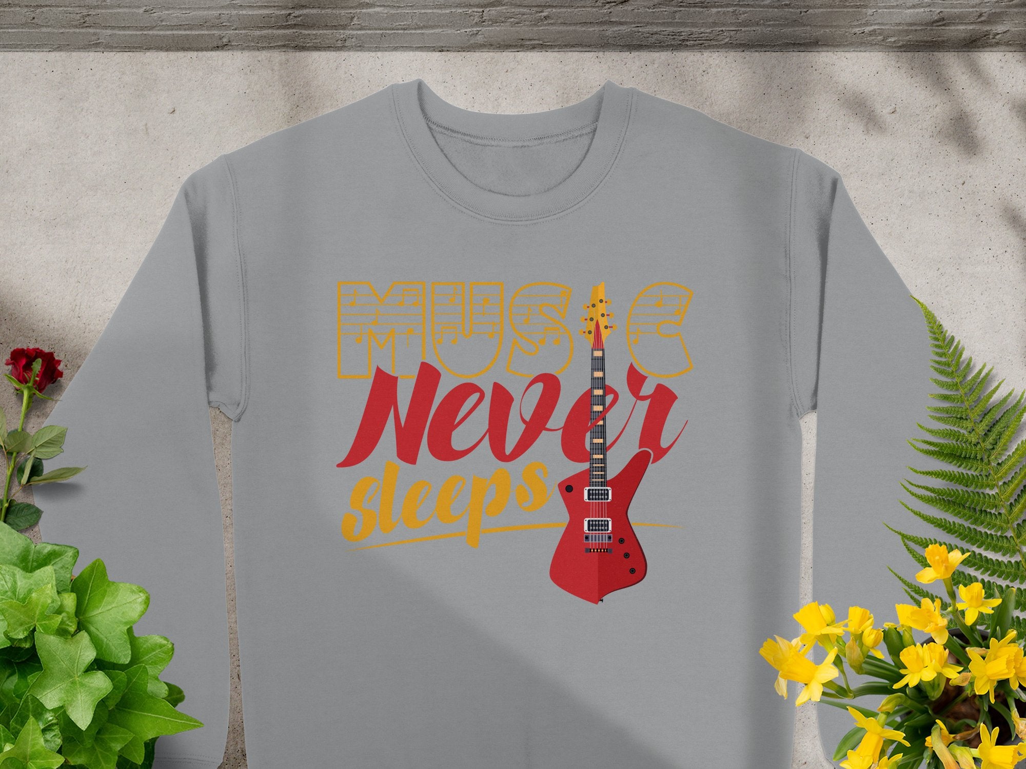 The Garment Graphics sweatshirt showcases Music Never Sleeps in bold red and yellow, with a vivid red guitar graphic. Ideal for music lovers, it’s beautifully set against green plants and yellow flowers on a light gray backdrop.