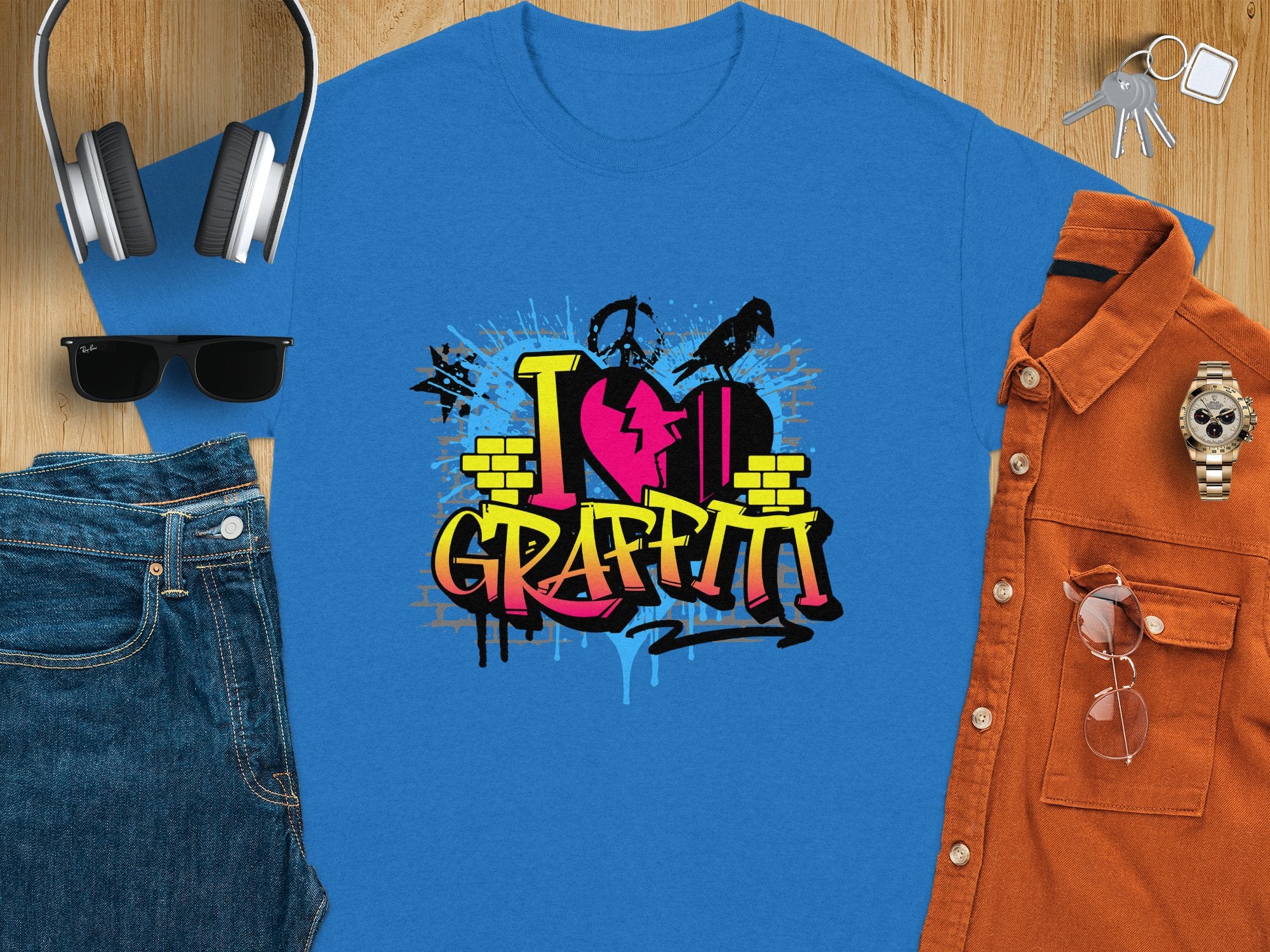 A blue t-shirt from Garment Graphics features a vivid Graffiti Love T-Shirts design with a pink heart and spray paint effect. Perfectly paired with headphones, sunglasses, a brown shirt, watch, jeans, and keys on a wooden surface, it embodies street art style.