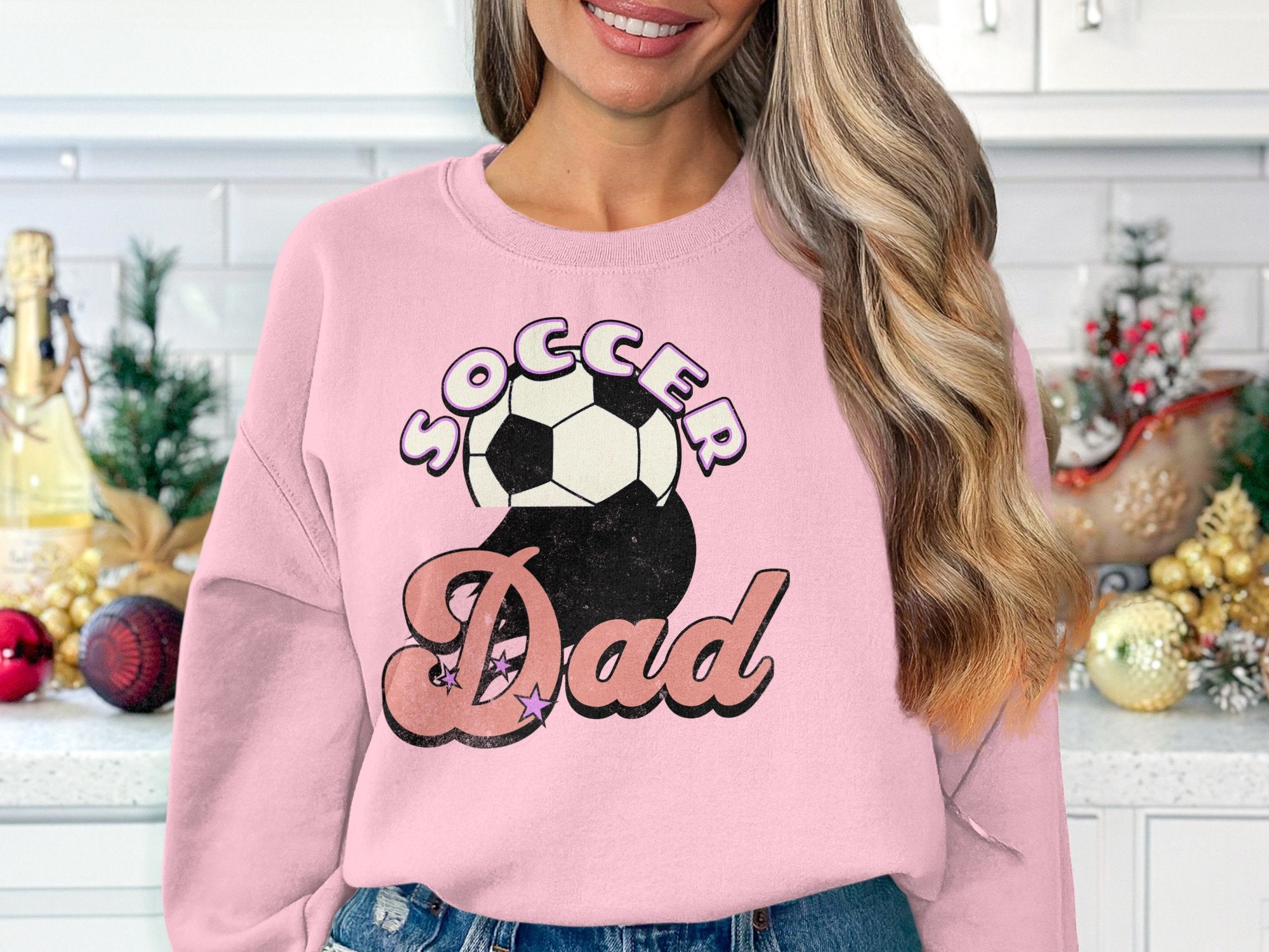 In a festive kitchen, a woman smiles in a Garment Graphics pink sweatshirt featuring Soccer Dad and a vintage soccer ball design. Her long hair cascades in loose waves amidst holiday-themed decorations.