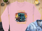 A pink graphic sweatshirt by Garment Graphics showcases a bold truck design and the phrase Keep On Trucking. This striking piece is displayed on a wooden surface next to a plant, latte, and denim shirt.