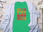 Garment Graphics green t-shirt, featuring Born to be a Musician in orange and white with drummer and guitarist silhouettes, is perfect for music lovers. Its styled with a gray cardigan and white flowers on a wooden surface.