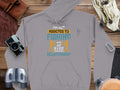 A Garment Graphics gray hoodie with Im not addicted to fishing, we are just in a very committed relationship in white, yellow, and blue adorns a wooden surface with travel items. Its classic fit makes it ideal for fishing enthusiasts, complemented by the camera and binoculars setting.