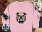 On a wooden surface, a Garment Graphics pink sweatshirt with a bulldog graphic and spiked collar is displayed. Nearby are a camera, binoculars, leather strap, pocket watch, and bag. This sweatshirt is available in unisex sizing.