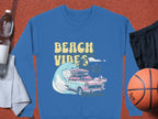 Garment Graphics blue sweatshirt showcases a vintage car, surfboards, a big wave, palm trees, and BEACH VIBES in yellow. This retro road trip design stands out on a textured brown background and pairs well with basketball gear like black shorts and a water bottle.
