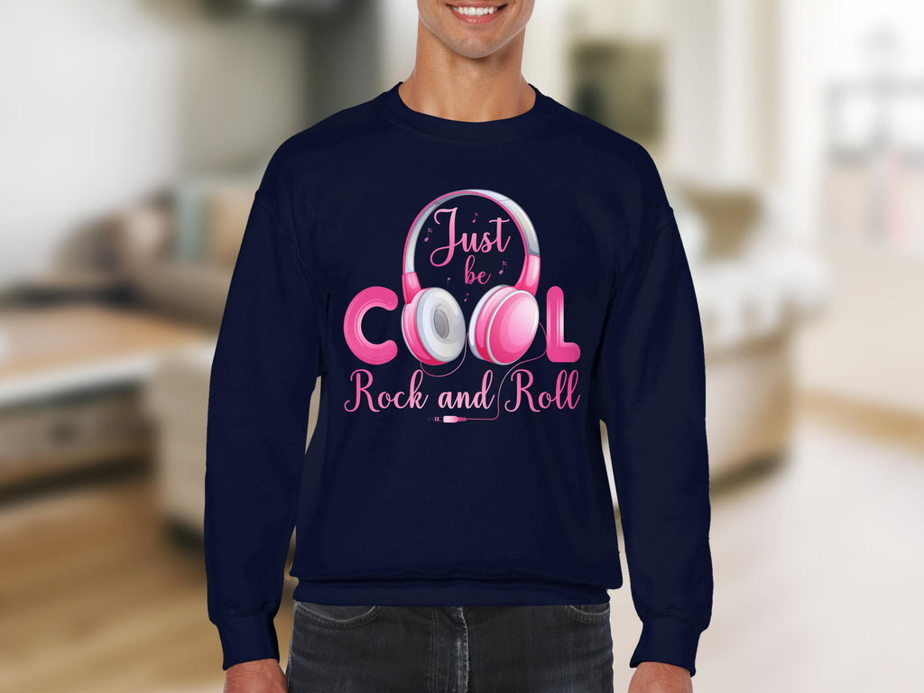 A person wears a navy blue Garment Graphics sweatshirt with Just be Cool Rock and Roll in pink and white. Fashion lovers will appreciate its design, with pink headphones around Cool, set against a blurred indoor backdrop thats ideal for music enthusiasts.