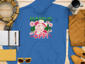 Garment Graphics presents a blue graphic hoodie showcasing a cowboy-hatted figure surrounded by cacti and flowers. The bold HANDS OFF! statement stands out on wooden attire, paired with a scarf, notebook, pen, yellow clutch, sunglasses, and coffee cup for an adventurous look.