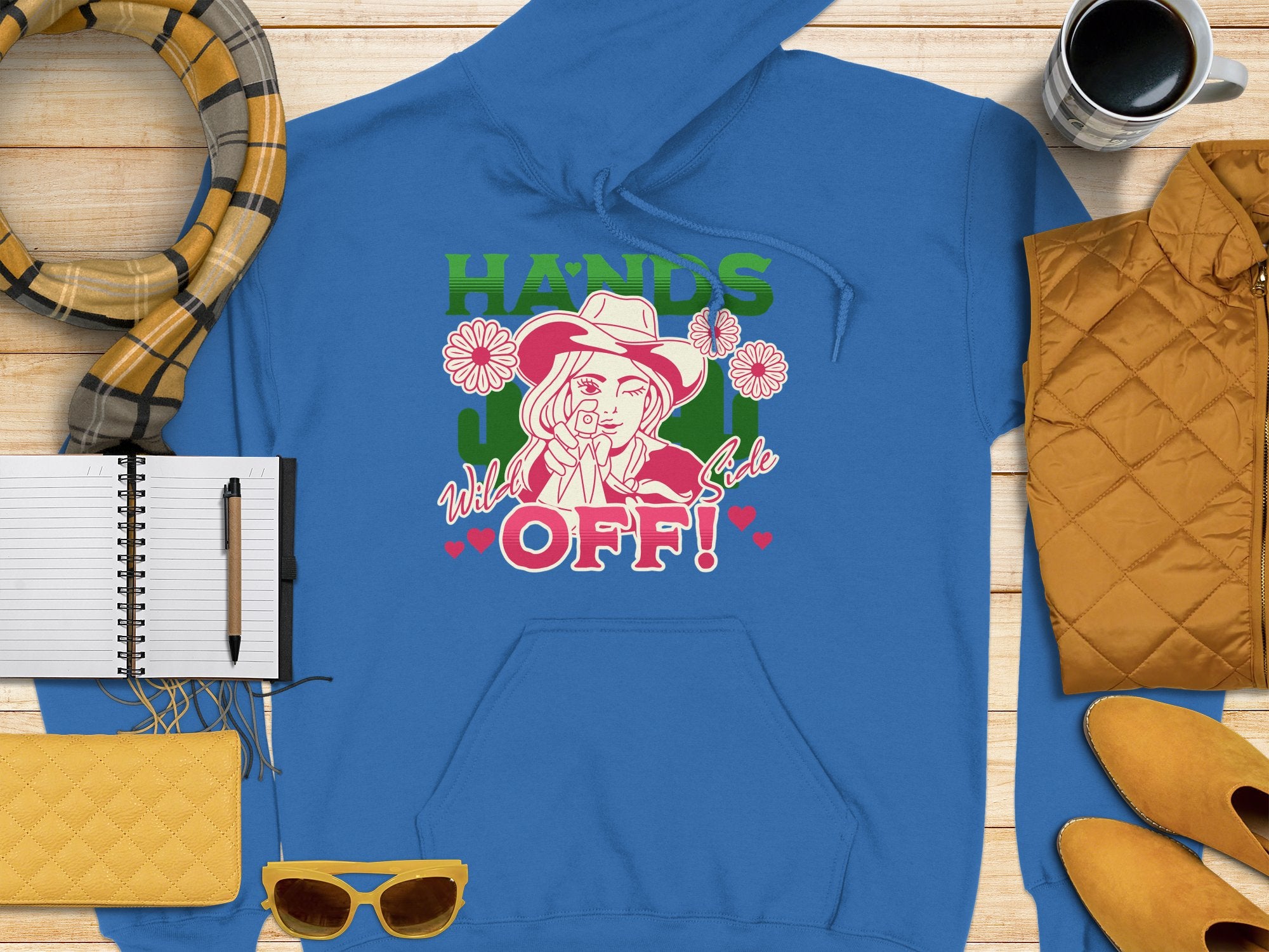 Garment Graphics presents a blue graphic hoodie showcasing a cowboy-hatted figure surrounded by cacti and flowers. The bold HANDS OFF! statement stands out on wooden attire, paired with a scarf, notebook, pen, yellow clutch, sunglasses, and coffee cup for an adventurous look.