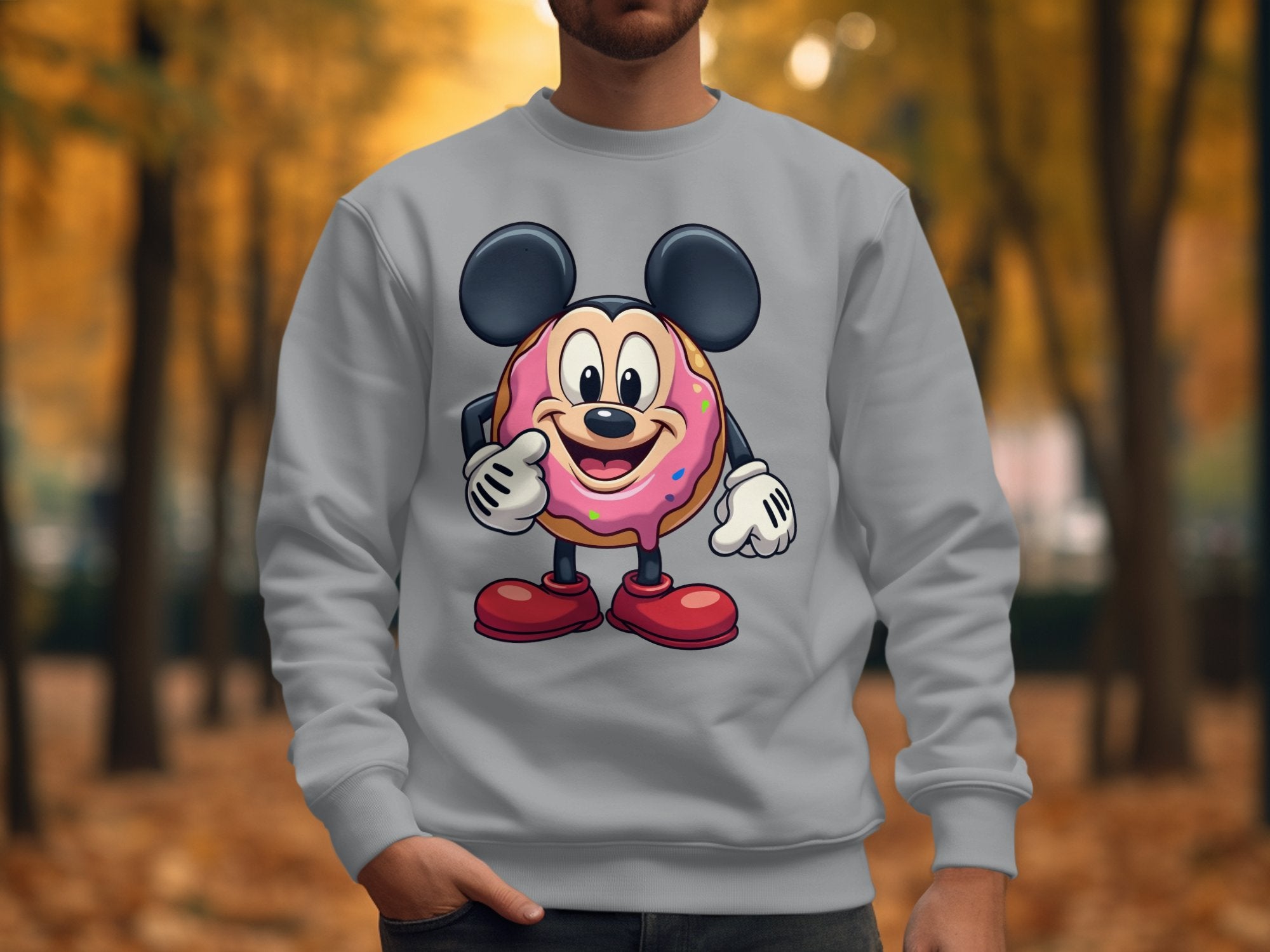 Sporting a quirky Garment Graphics sweatshirt depicting a cute cartoon character with large ears and red shoes, holding a pink frosted donut with sprinkles on an outdoor path lined with trees and fallen leaves, adding to the playful wardrobe vibe.